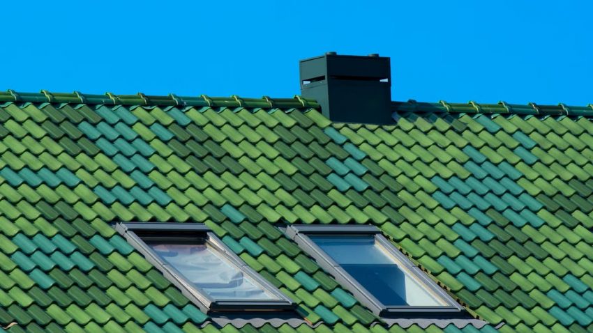 A modern architectural roof with green tiles, skylights, and chimney against a blue sky.